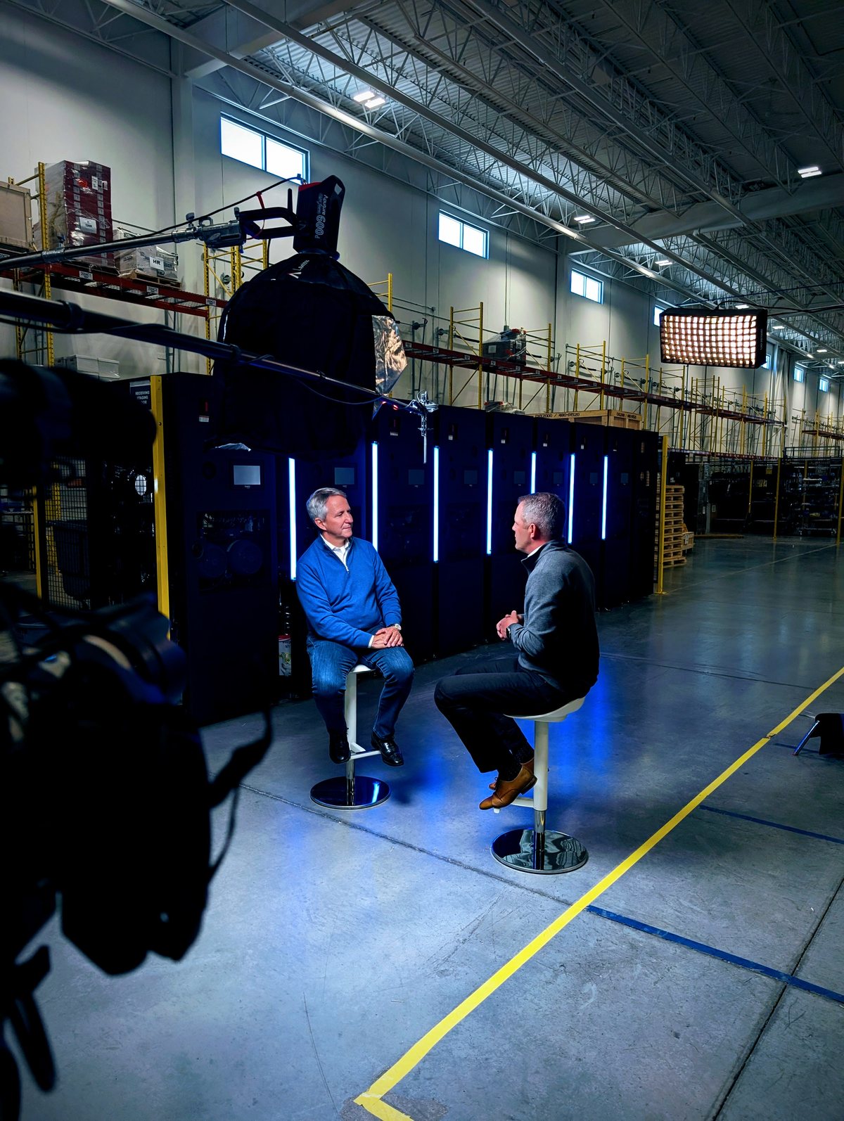 Two company leaders seated for a filmed interview on the finished blue-lit set inside the CoolIT facility, Calgary