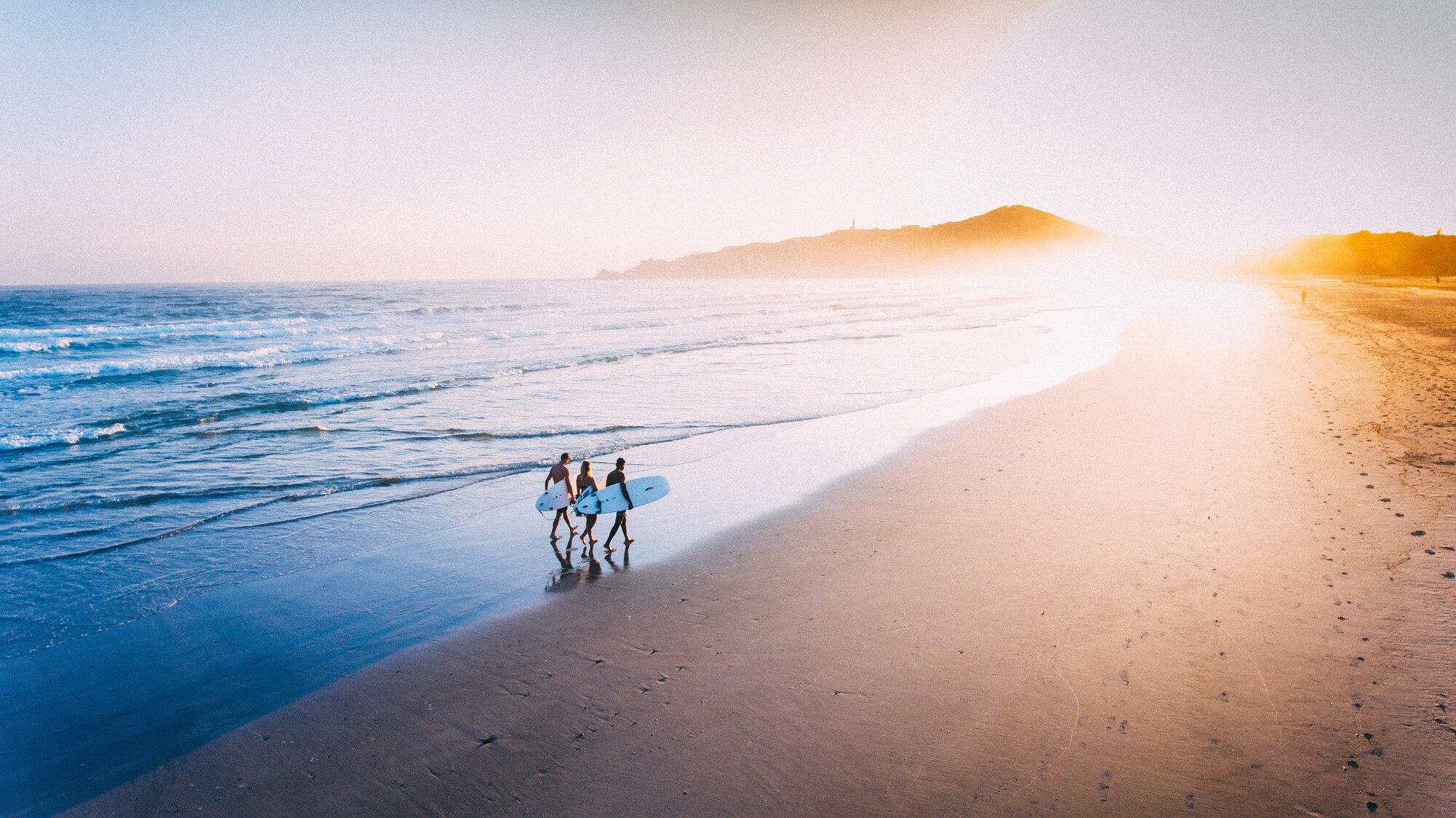 Two surfers walking a bright beach at sunrise, cinematic aerial drone shot for commercial production
